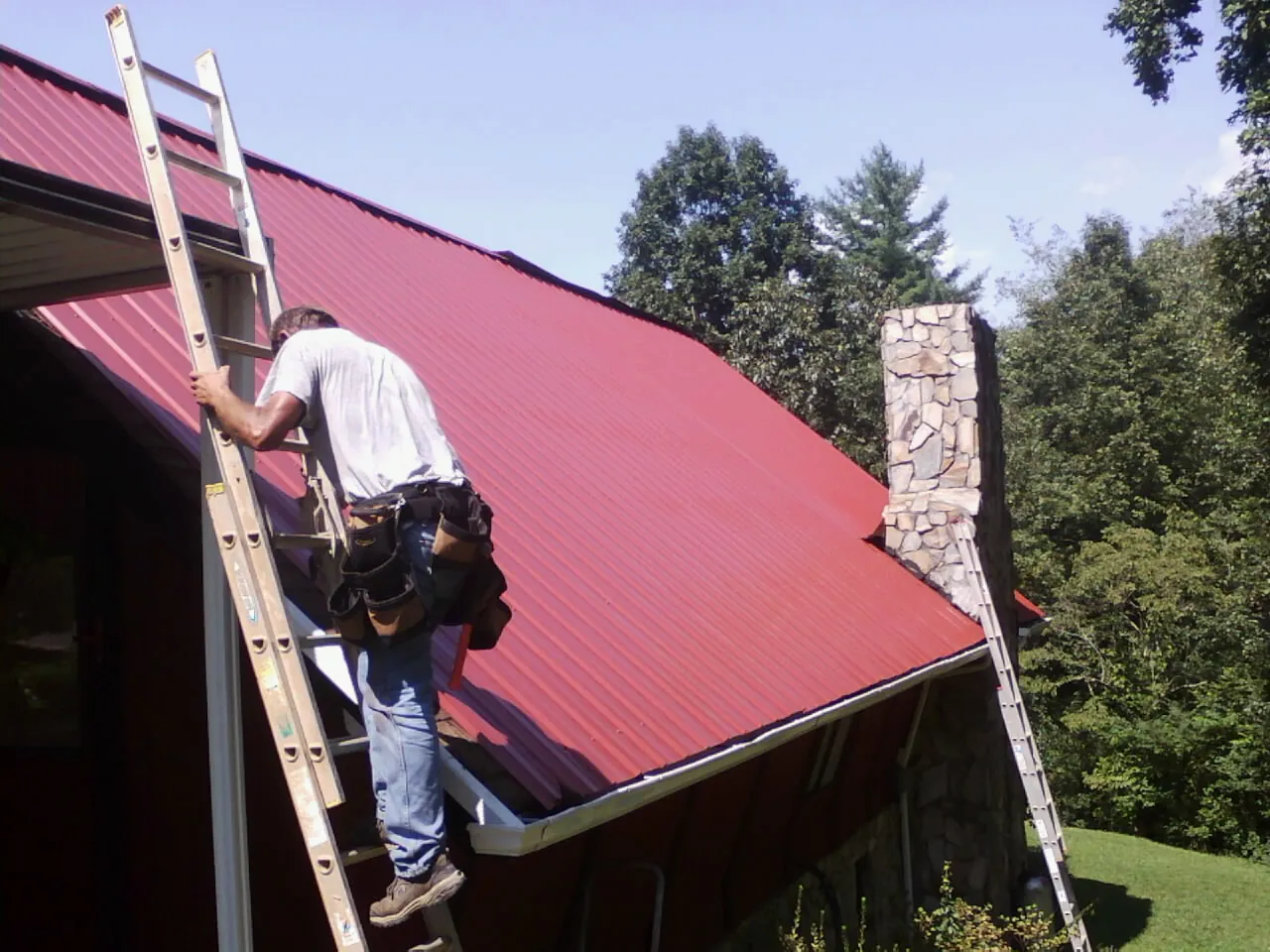 Roofer on a ladder beside a red metal roof and stone chimney — Soffit & Fascia Repair by Roofing Brothers in Pine Beach